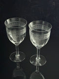 Two clear cut-crystal wine glasses with etched geometric patterns on the bowls, standing on a dark reflective surface against a dark background.