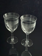 Two clear cut-crystal wine glasses with etched geometric patterns on the bowls, standing on a dark reflective surface against a dark background.