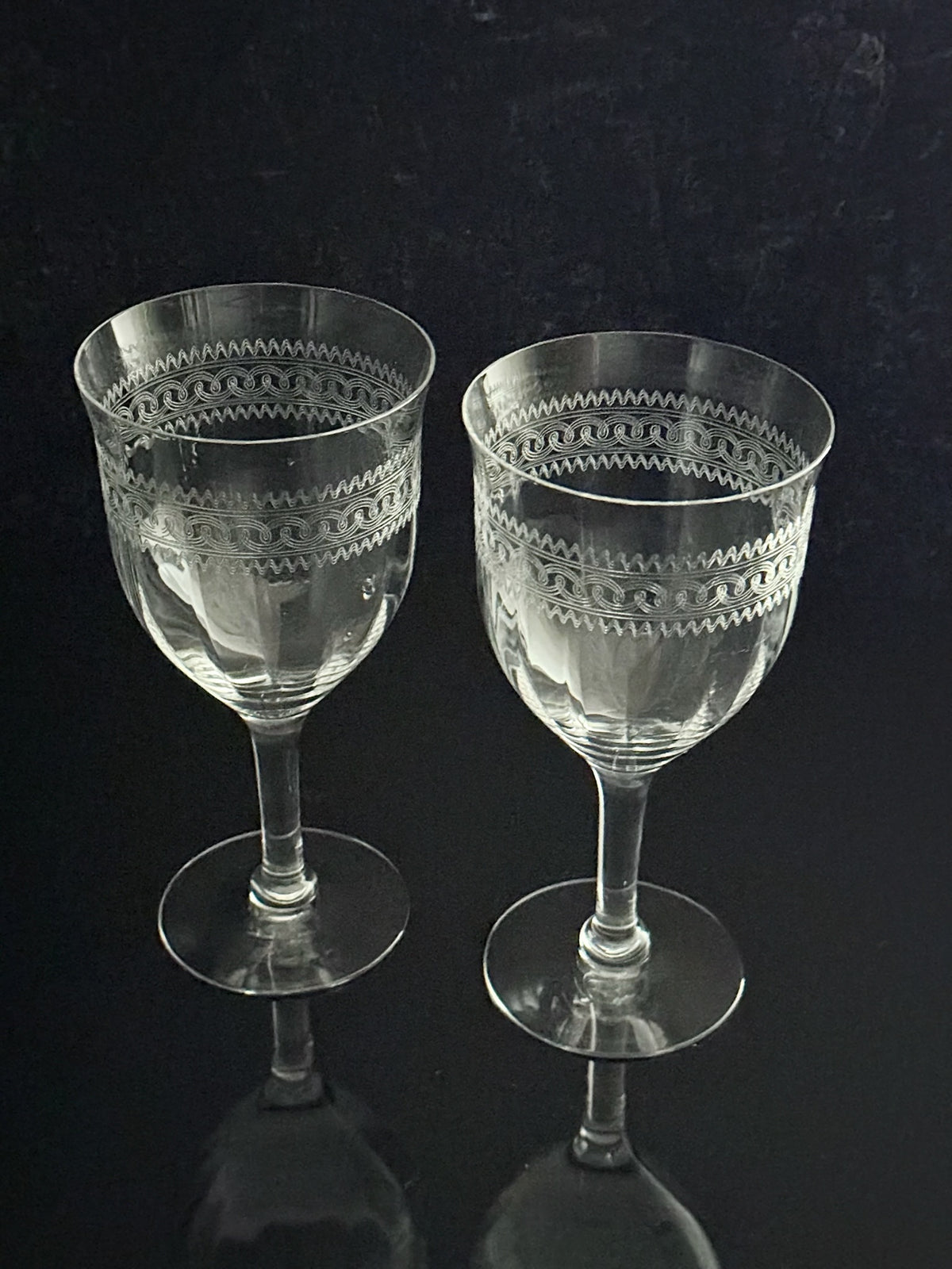 Two clear cut-crystal wine glasses with etched geometric patterns on the bowls, standing on a dark reflective surface against a dark background.