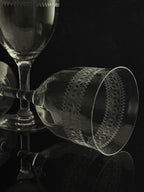A clear etched glass goblet with geometric patterns, shown lying on its side on a reflective black surface, with other glassware blurred in the background.