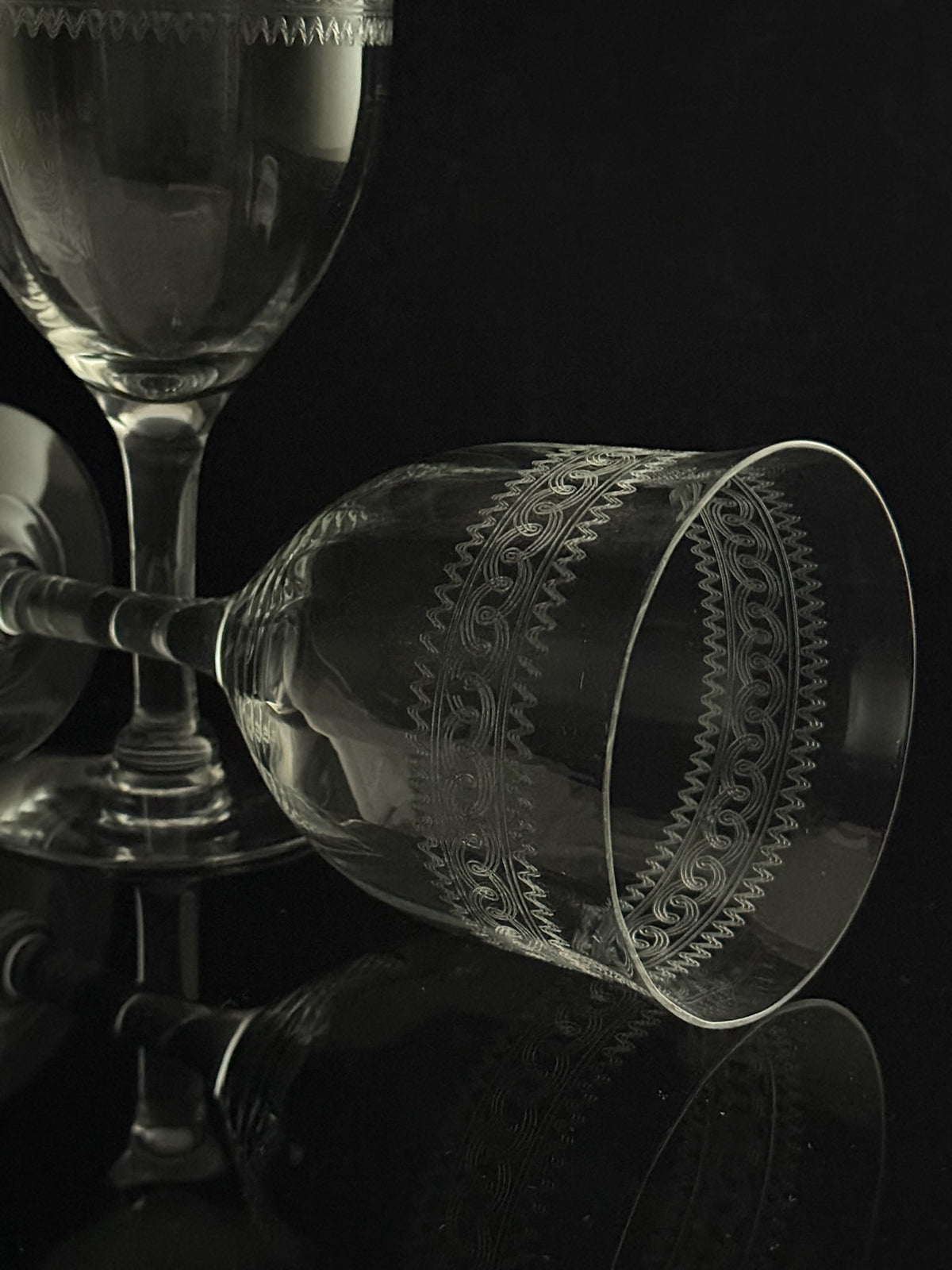 A clear etched glass goblet with geometric patterns, shown lying on its side on a reflective black surface, with other glassware blurred in the background.