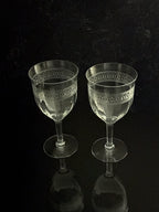 Two clear cut-crystal wine glasses with geometric patterns on the bowls, standing on a reflective black surface against a dark background.