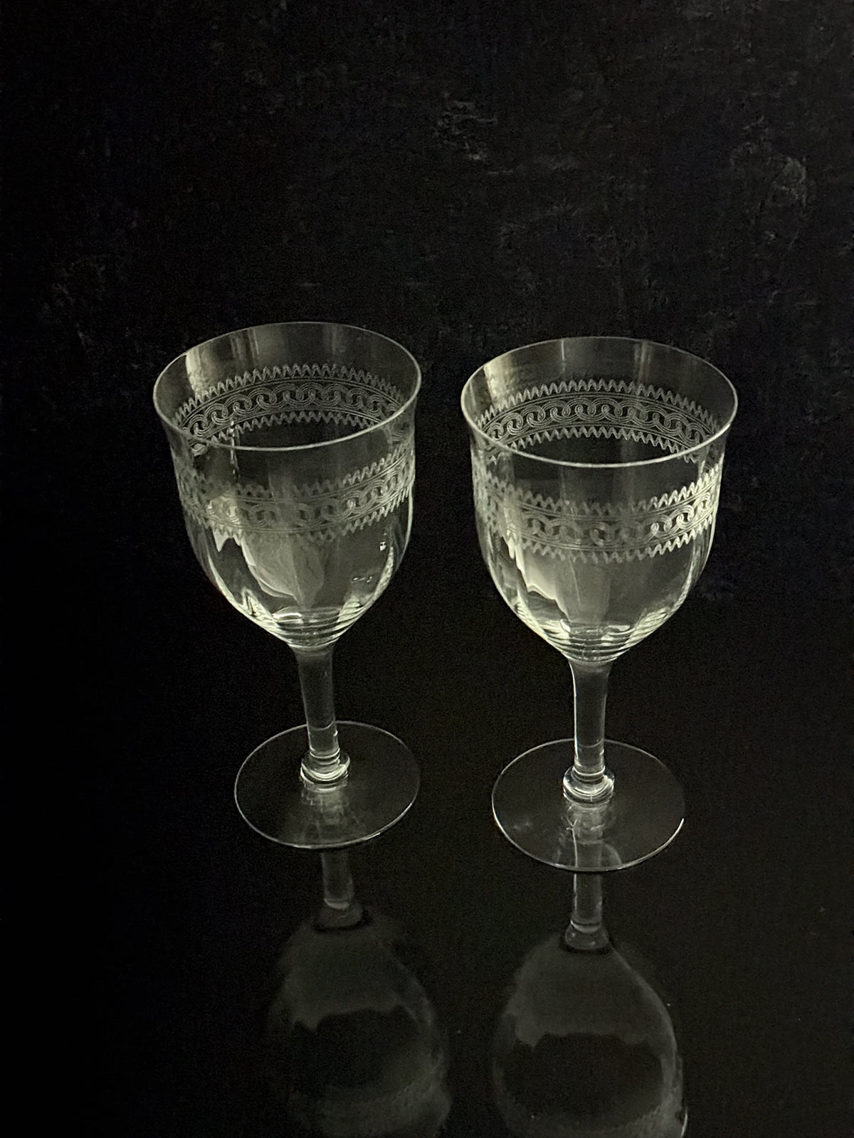 Two clear cut-crystal wine glasses with geometric patterns on the bowls, standing on a reflective black surface against a dark background.
