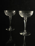 Two clear cut-crystal coupe glasses with etched leaf patterns, displayed on a reflective black surface against a dark background.