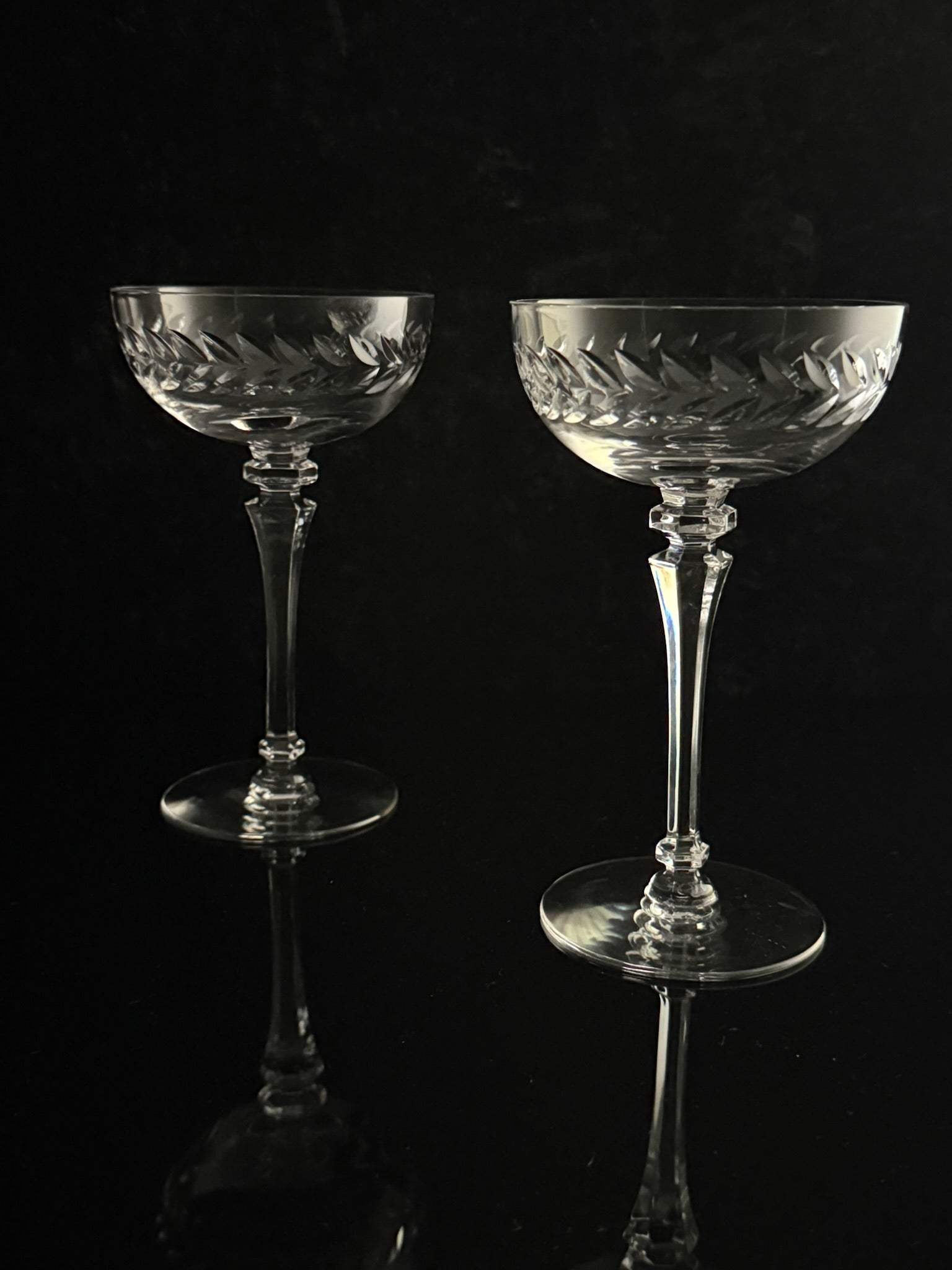 Two clear cut-crystal coupe glasses with etched leaf patterns, displayed on a reflective black surface against a dark background.