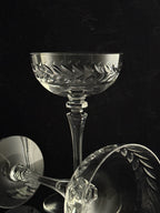 Clear cut-crystal coupe glasses with a leaf-patterned bowl, shown on a dark black background with one glass standing upright and two cropped reflections/angles in the foreground.