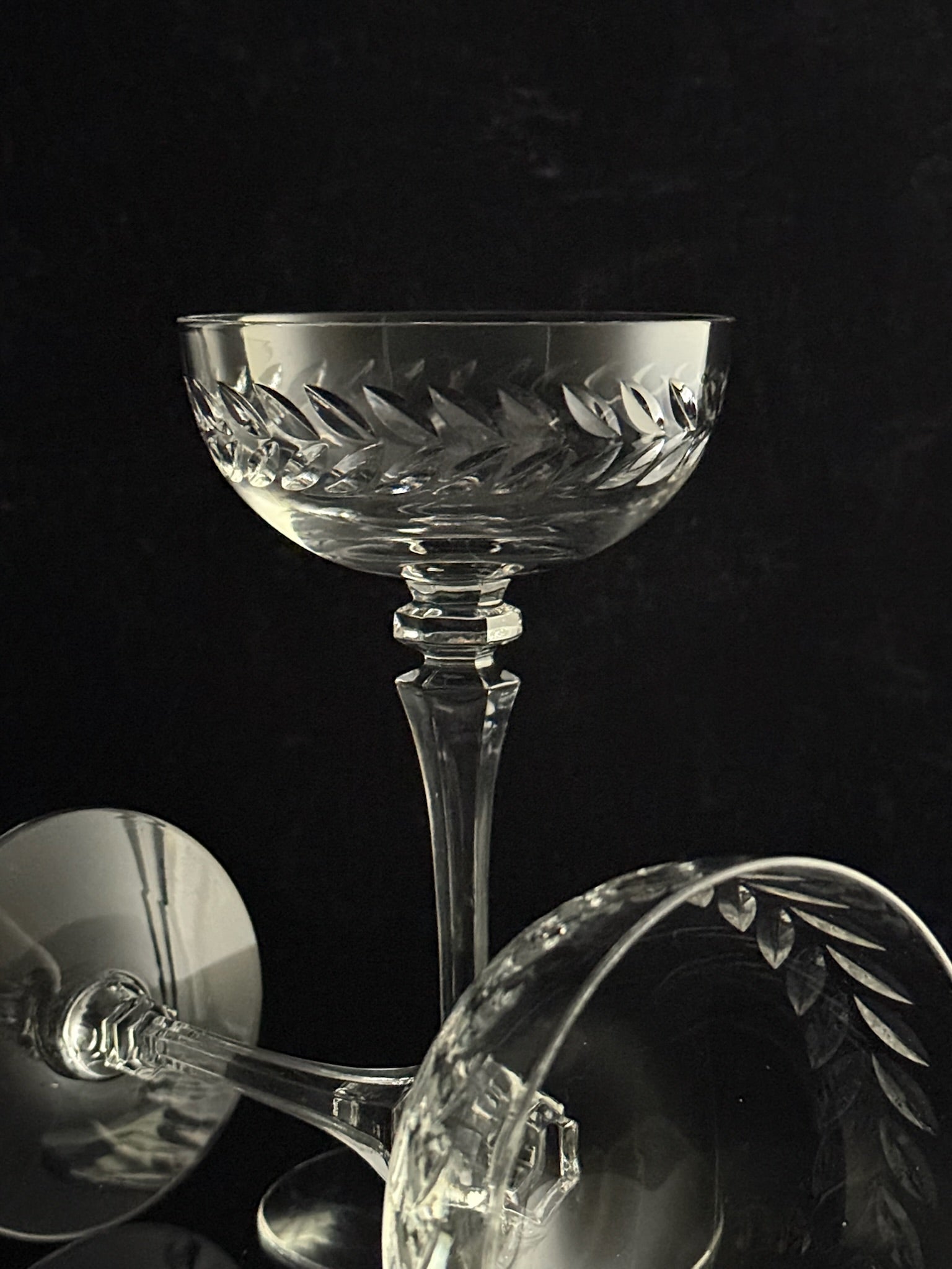 Clear cut-crystal coupe glasses with a leaf-patterned bowl, shown on a dark black background with one glass standing upright and two cropped reflections/angles in the foreground.