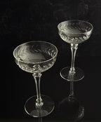 Two clear cut-crystal coupe glasses with etched floral patterns, displayed on a reflective black surface against a dark background.