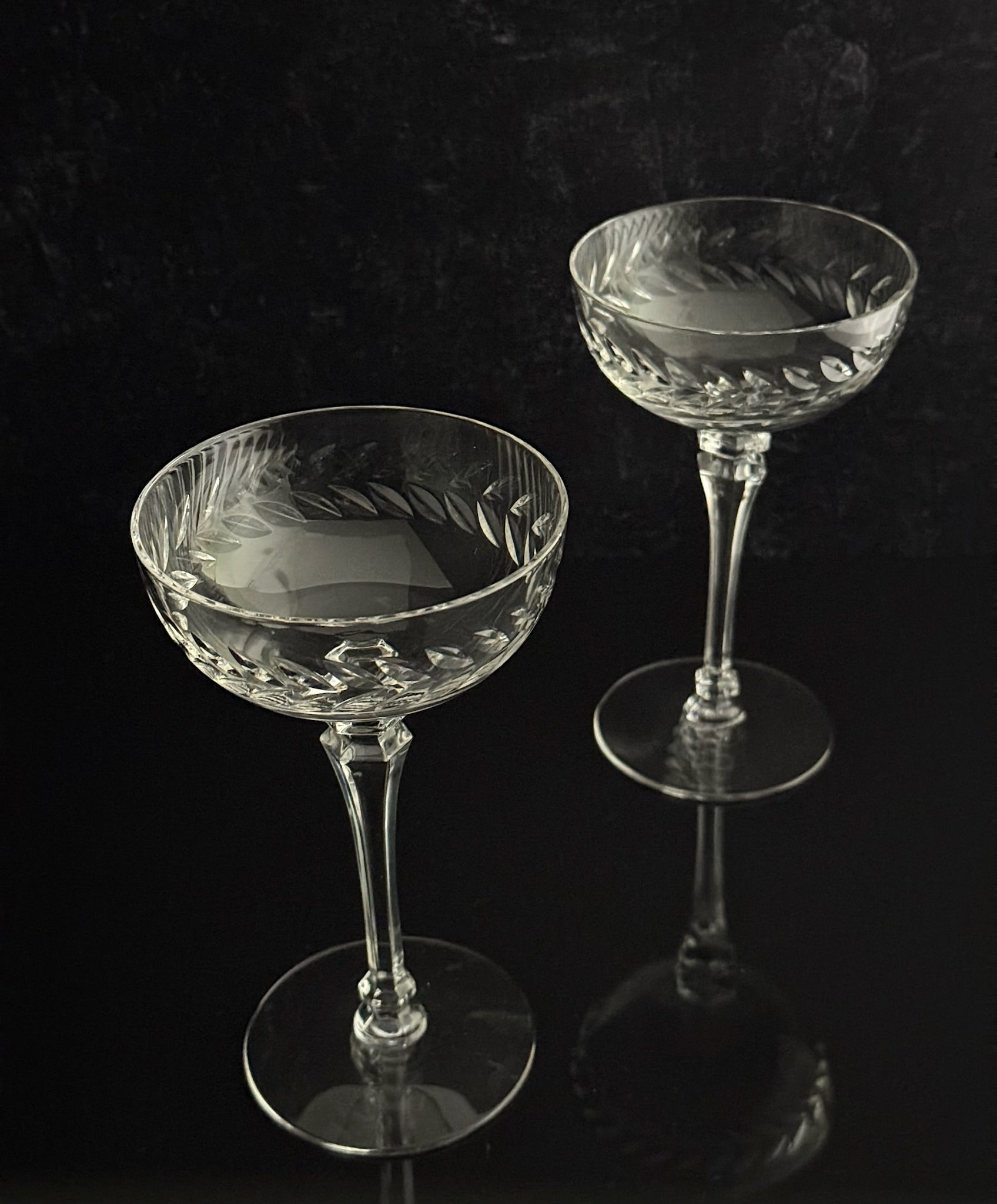 Two clear cut-crystal coupe glasses with etched floral patterns, displayed on a reflective black surface against a dark background.