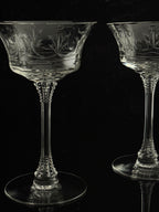 Two clear crystal coupe glasses with etched floral patterns, displayed on a reflective black surface against a dark background.
