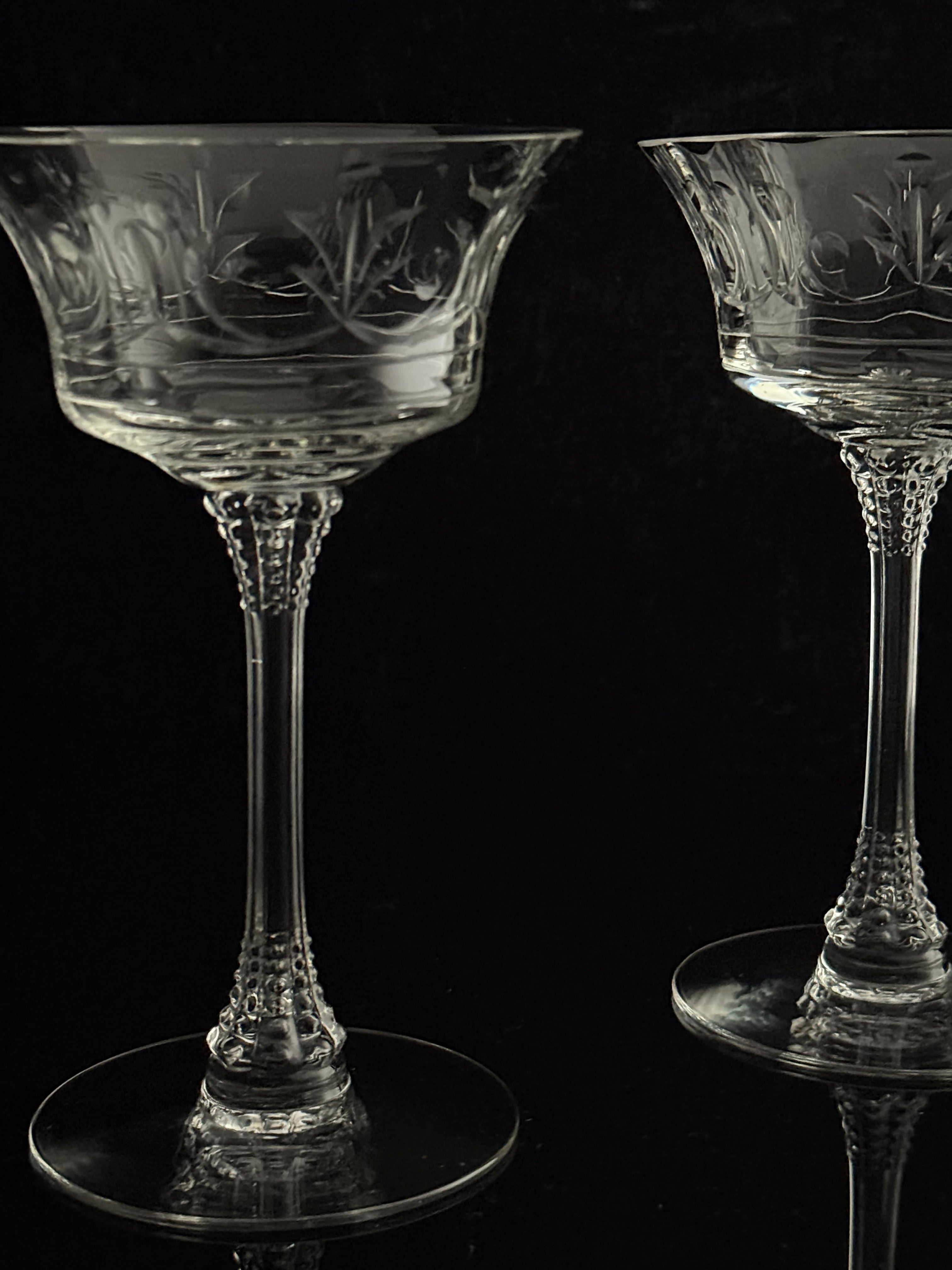 Two clear crystal coupe glasses with etched floral patterns, displayed on a reflective black surface against a dark background.