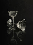 Two clear cut-crystal coupe glasses with etched floral patterns, one standing upright and the other lying on its side, set against a dark reflective black background.