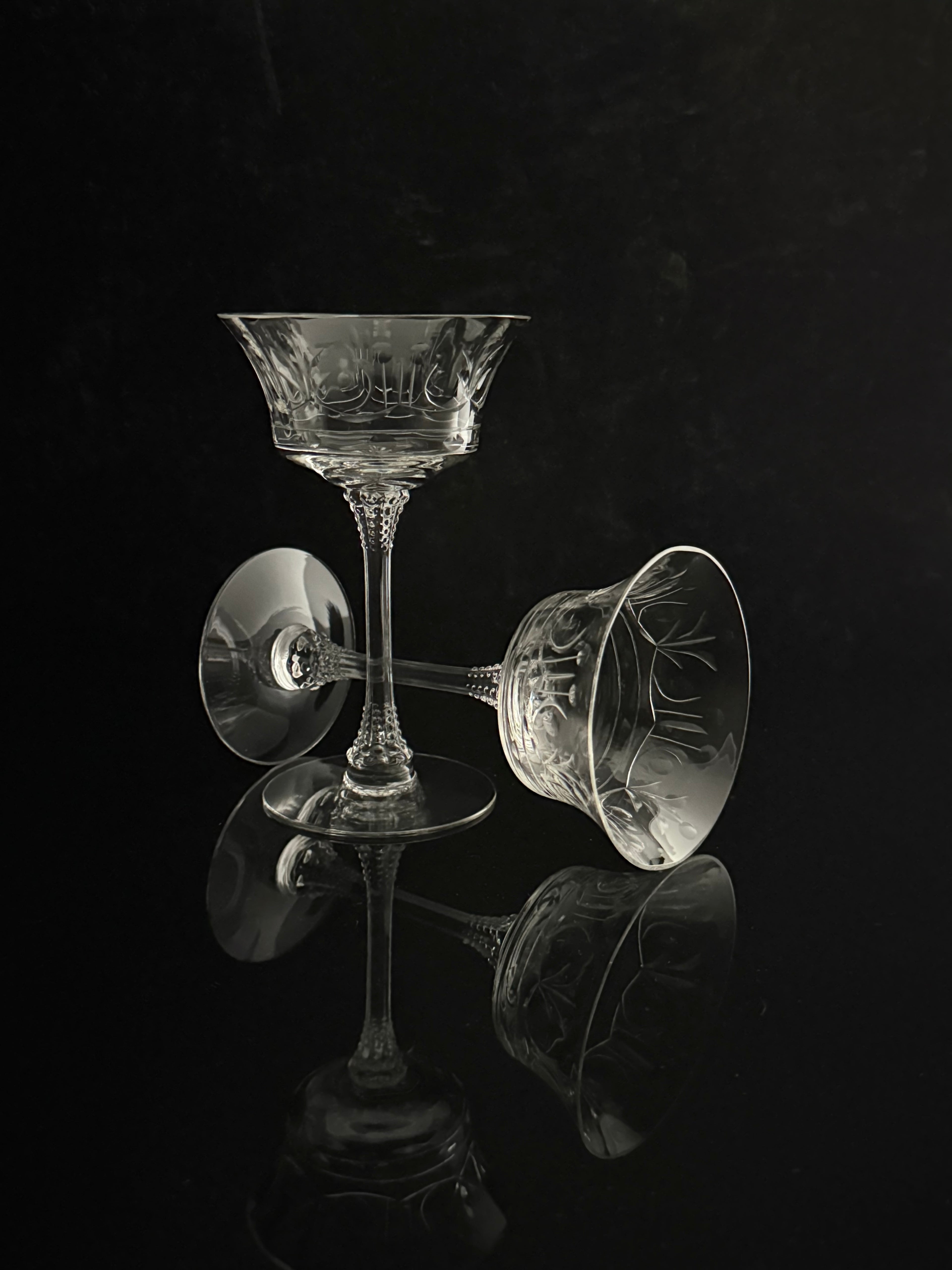 Two clear cut-crystal coupe glasses with etched floral patterns, one standing upright and the other lying on its side, set against a dark reflective black background.