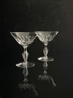 Two clear cut-crystal coupe glasses with elegant stems and wide bowls, set against a dark reflective background.