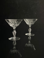 Two clear cut-crystal coupe glasses with faceted stems and circular bases, displayed on a reflective black surface against a dark background.