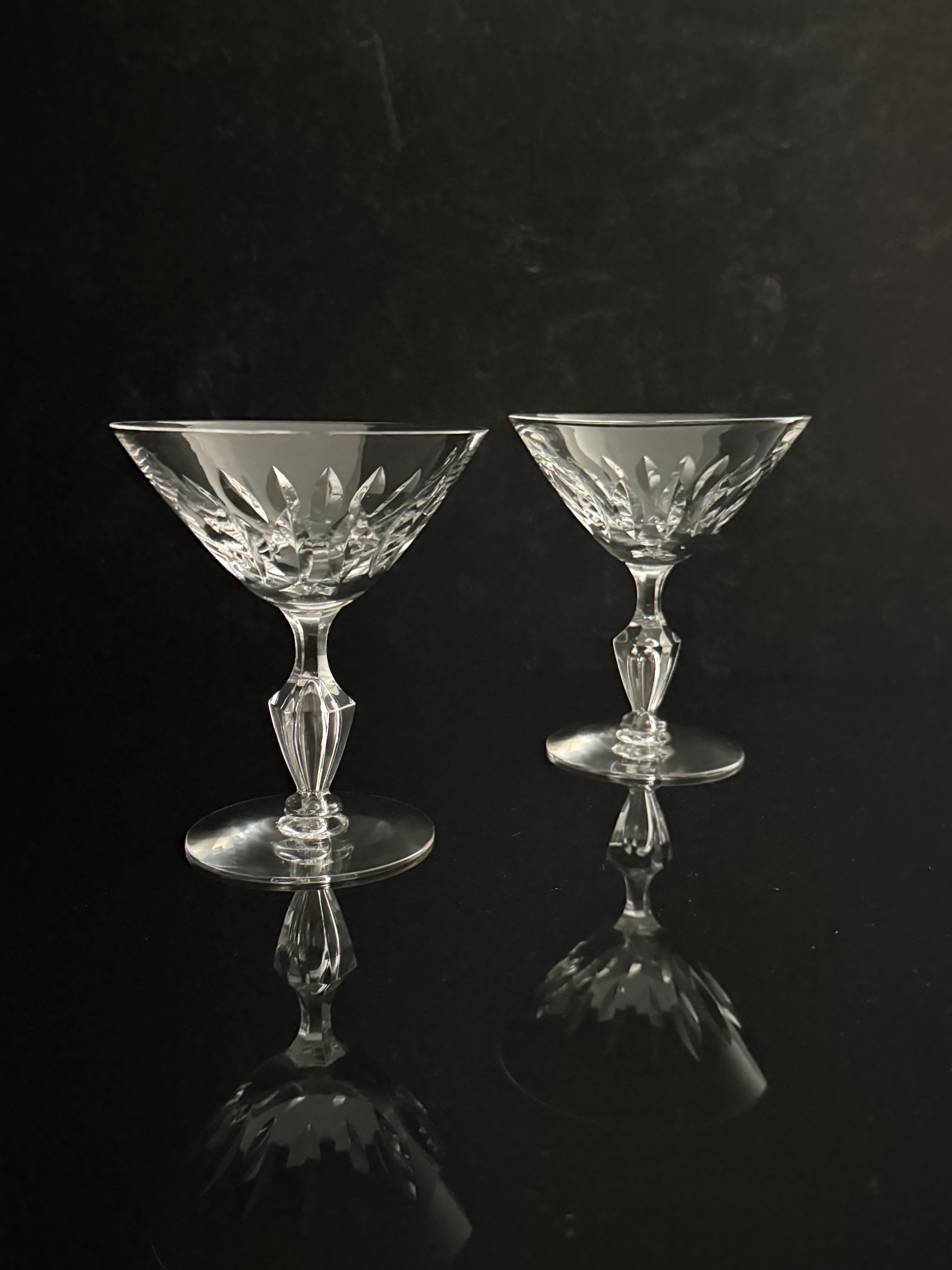 Two clear cut-crystal coupe glasses with faceted stems and circular bases, displayed on a reflective black surface against a dark background.