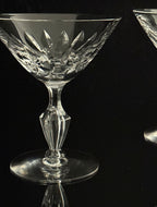 A clear cut-crystal coupe glass with a faceted stem and circular base, photographed against a black reflective background. Part of a second matching glass is visible at the right edge.