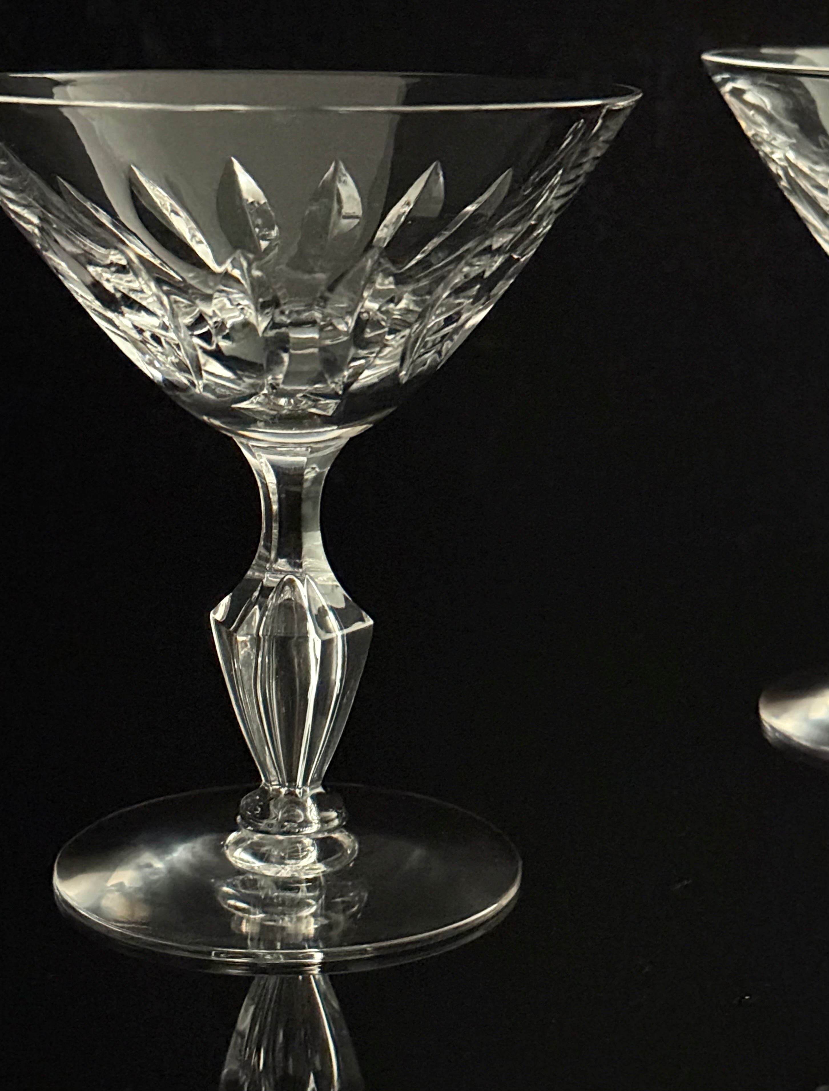 A clear cut-crystal coupe glass with a faceted stem and circular base, photographed against a black reflective background. Part of a second matching glass is visible at the right edge.