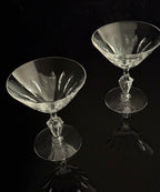 Two clear cut-crystal coupe glasses with faceted stems and circular bases, photographed on a glossy black reflective surface against a black background.