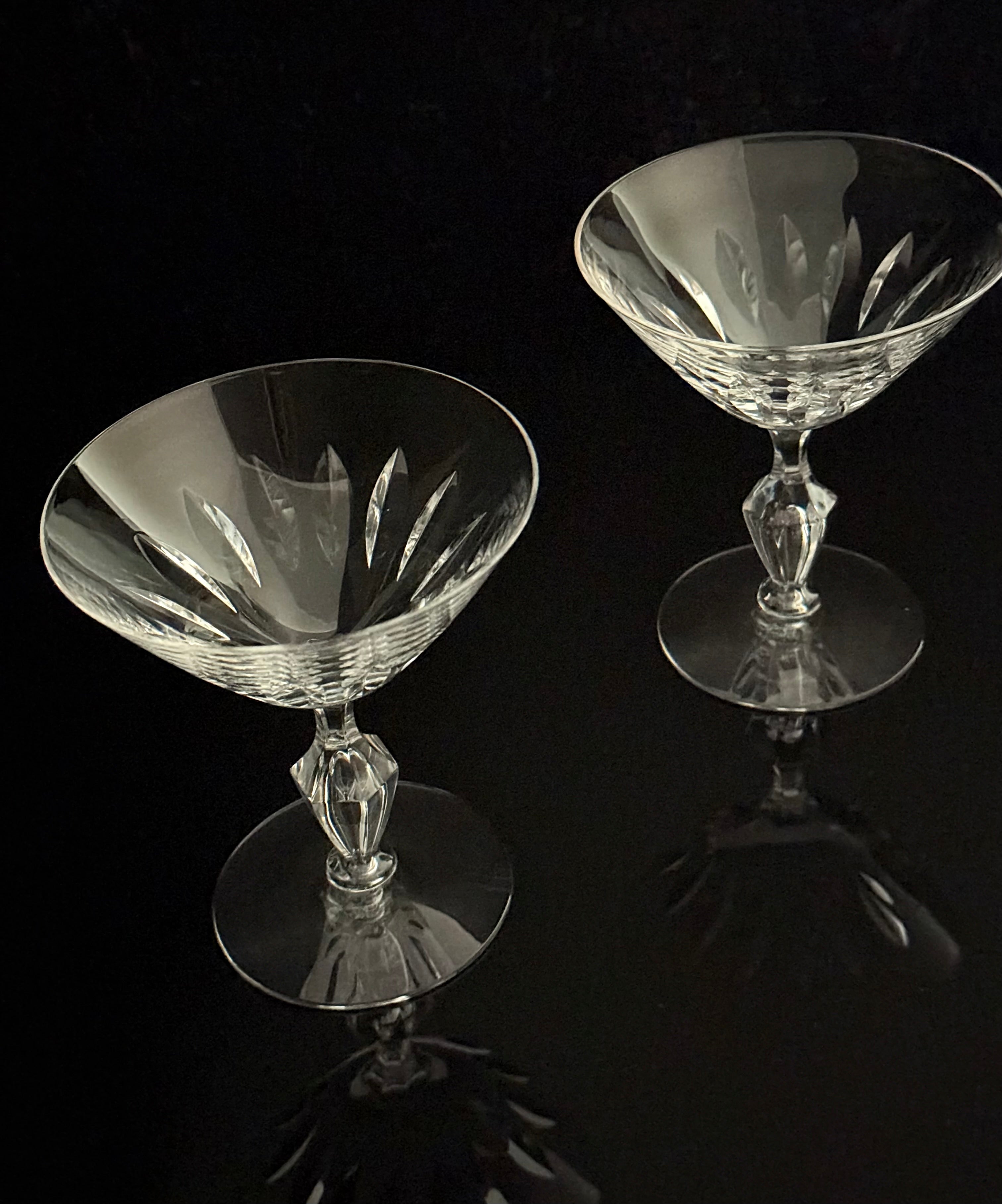 Two clear cut-crystal coupe glasses with faceted stems and circular bases, photographed on a glossy black reflective surface against a black background.