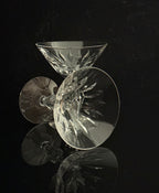 A set of clear cut-crystal cocktail glasses with intricate faceted patterns, arranged on a reflective black surface against a dark background.
