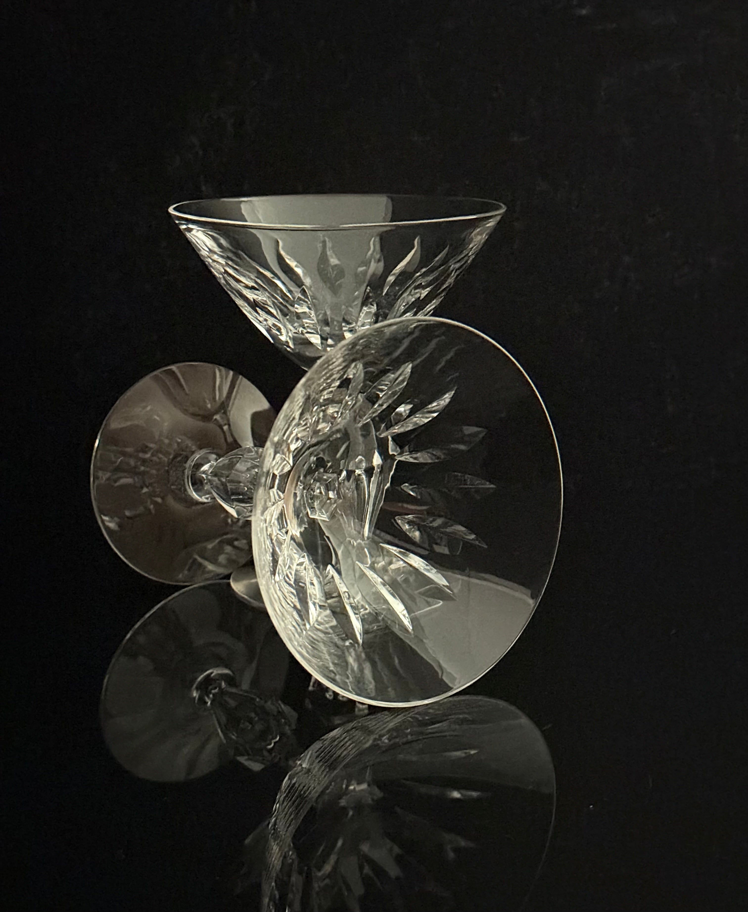 A set of clear cut-crystal cocktail glasses with intricate faceted patterns, arranged on a reflective black surface against a dark background.