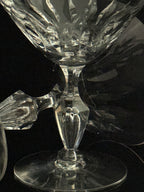 A close-up of a clear cut-crystal goblet or cocktail glass with a faceted stem and circular base, photographed against a dark reflective background.