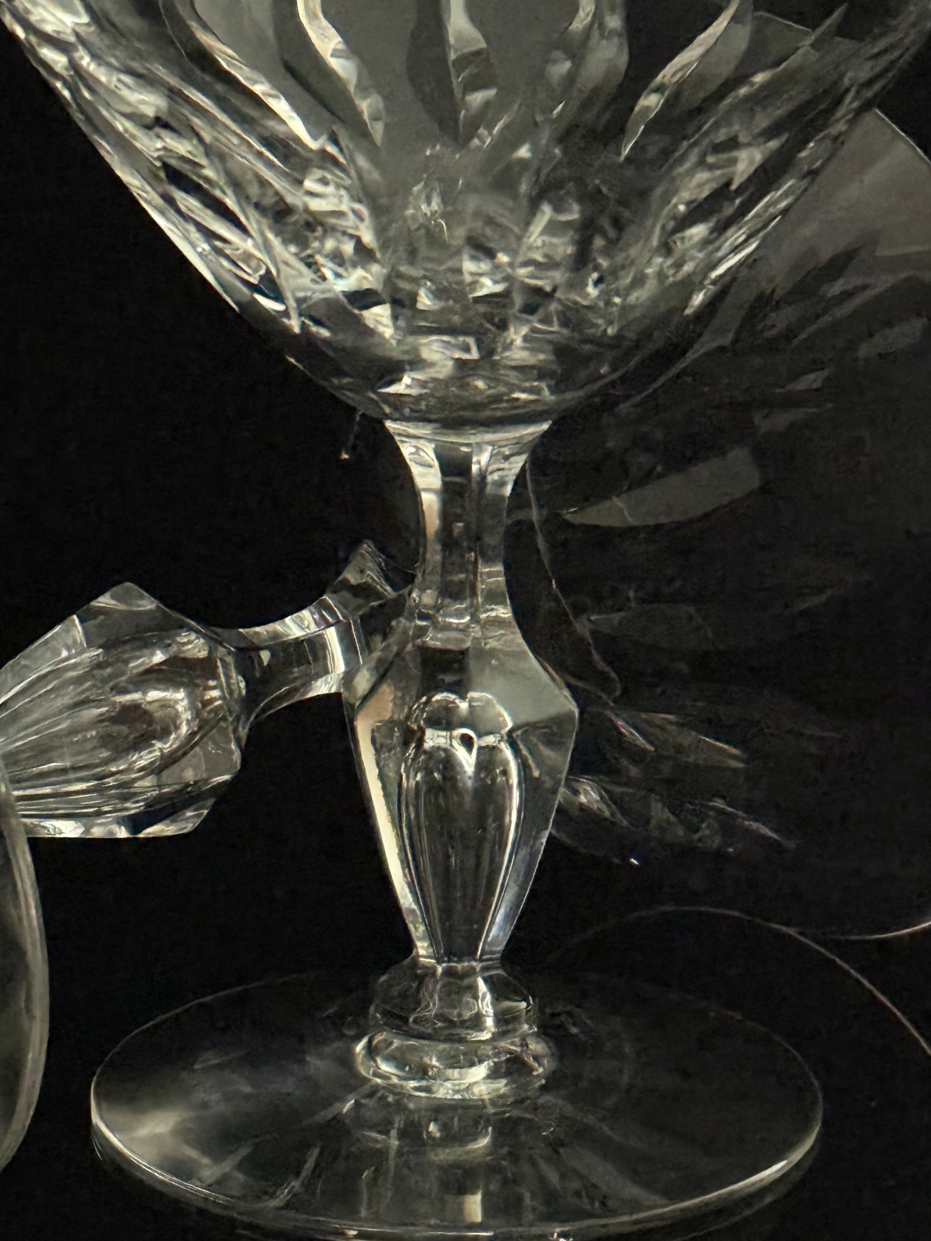 A close-up of a clear cut-crystal goblet or cocktail glass with a faceted stem and circular base, photographed against a dark reflective background.