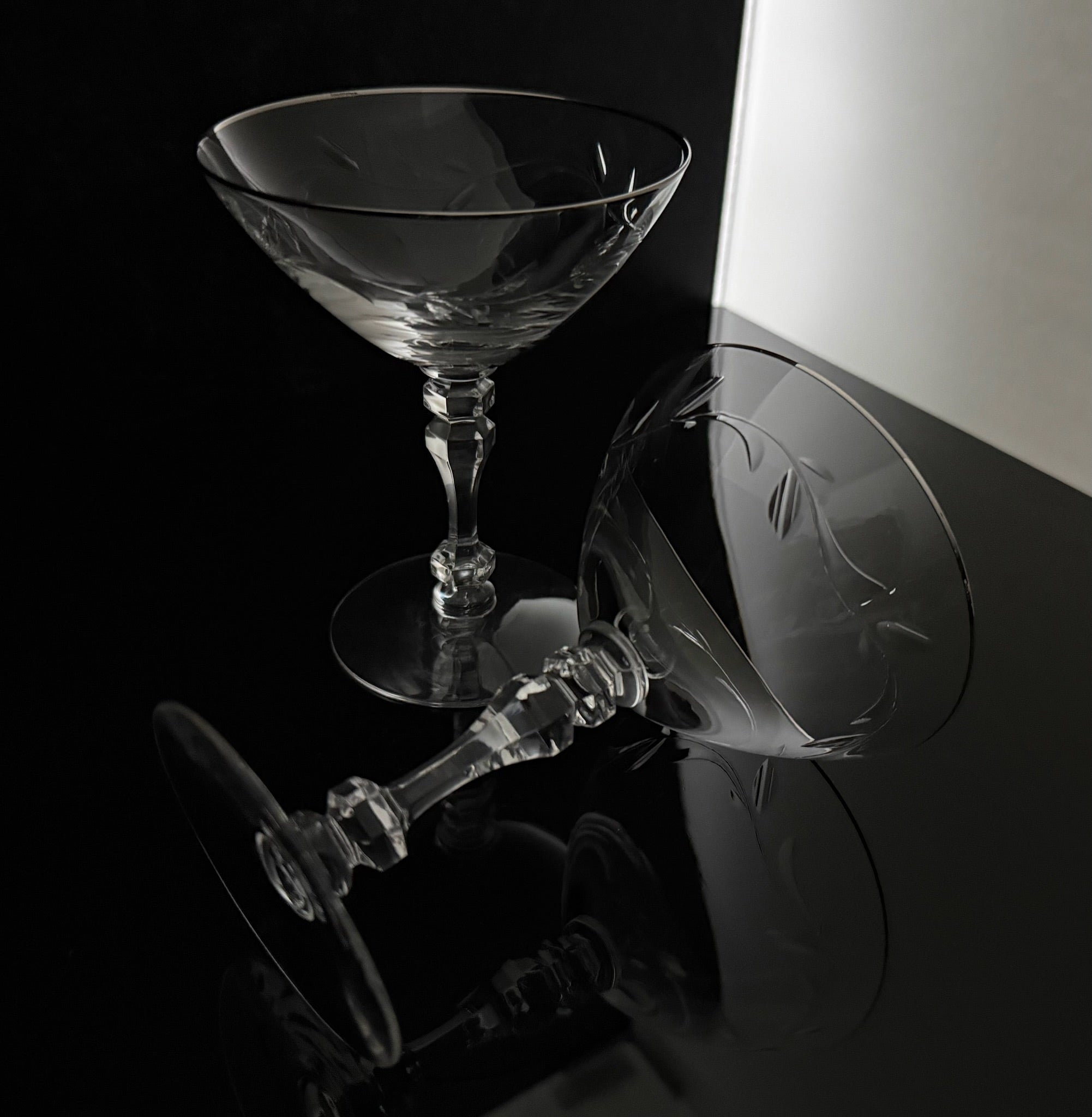 A clear cut-glass coupe-style cocktail glass shown on a glossy black surface, with one upright glass and one lying on its side, both reflecting in the dark. A white wall is visible in the background.