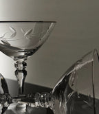 Clear crystal cocktail coupe glasses with etched leaf patterns, shown in a close-up studio arrangement against a neutral gray background.