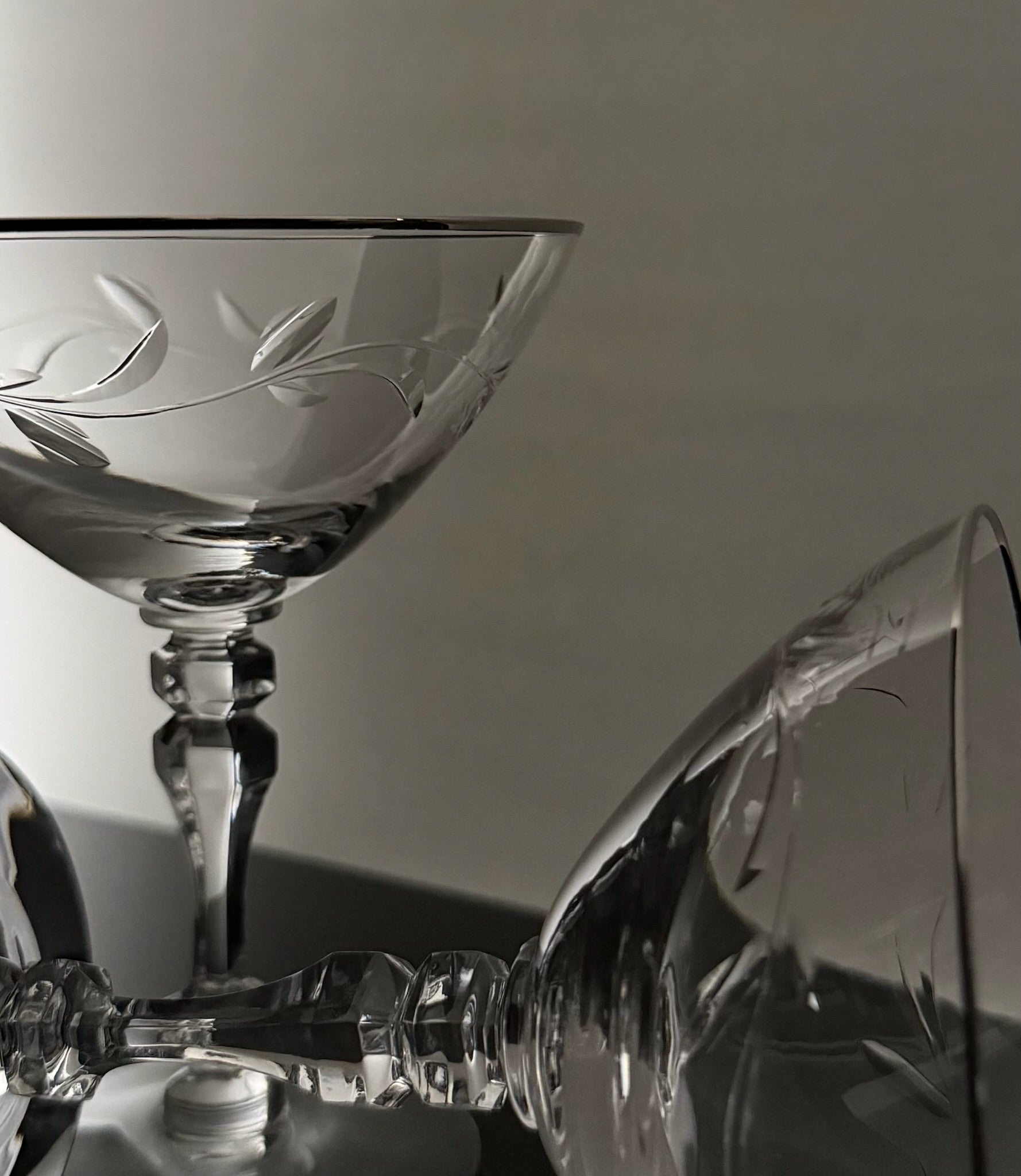 Clear crystal cocktail coupe glasses with etched leaf patterns, shown in a close-up studio arrangement against a neutral gray background.