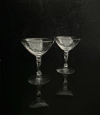Two clear cut-glass coupe glasses with faceted stems and circular bases, standing on a glossy black reflective surface against a dark black background.