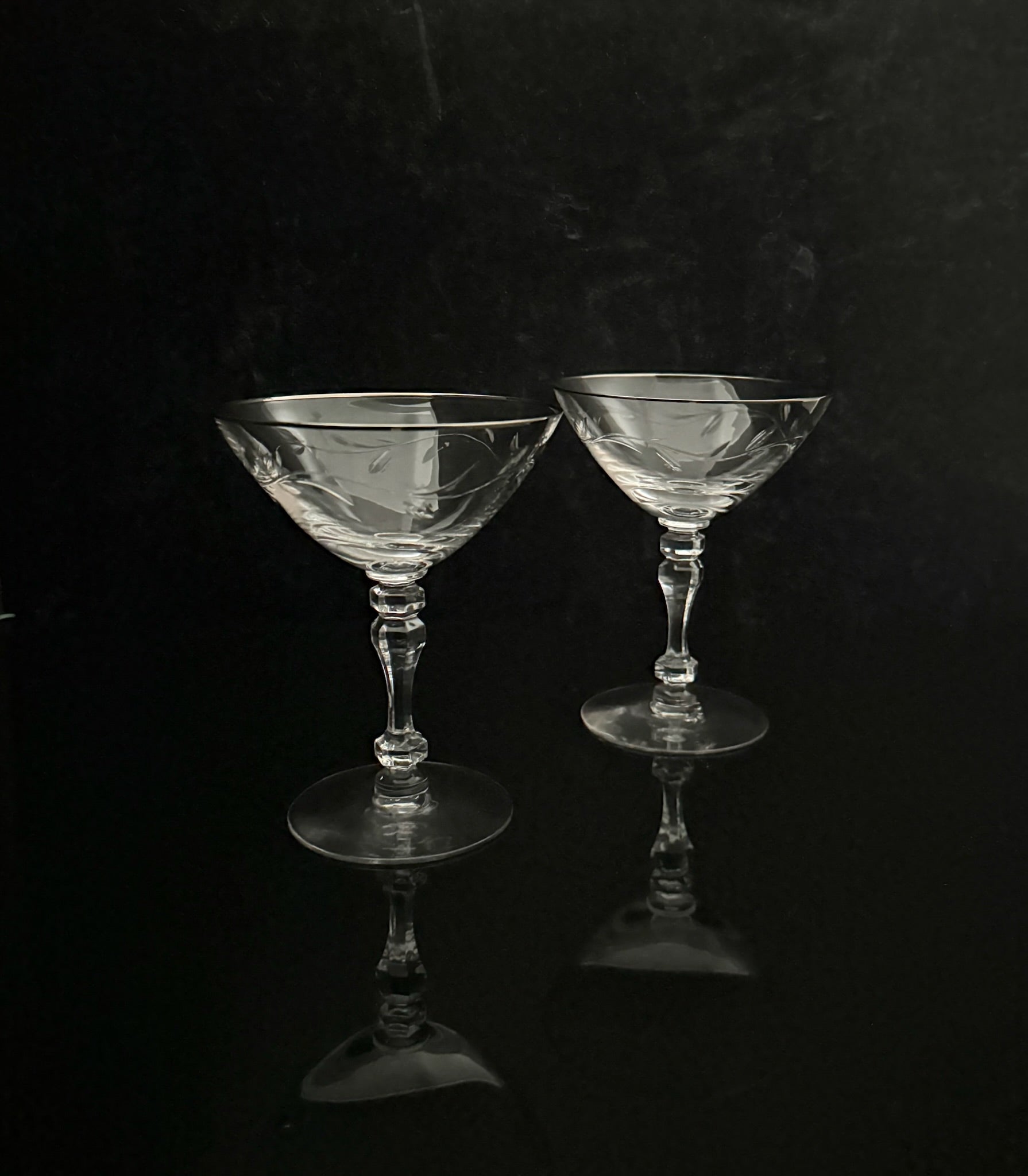 Two clear cut-glass coupe glasses with faceted stems and circular bases, standing on a glossy black reflective surface against a dark black background.