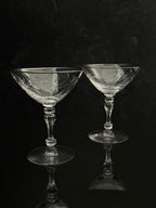 Two clear cut-glass coupe glasses with etched floral patterns, standing on a reflective black surface against a dark background.