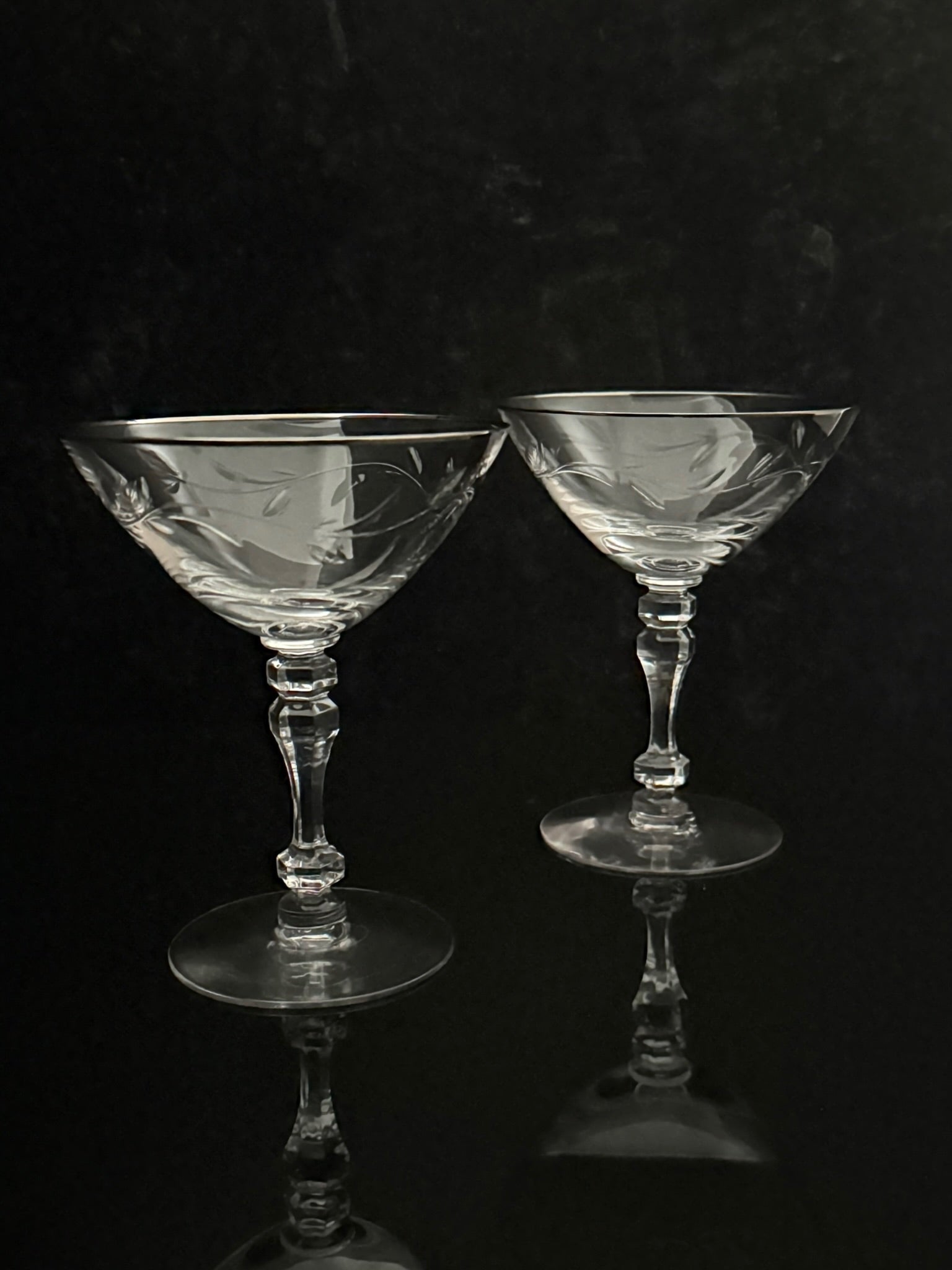 Two clear cut-glass coupe glasses with etched floral patterns, standing on a reflective black surface against a dark background.