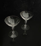 Two clear cut-glass coupe glasses with faceted stems and circular bases, photographed on a glossy black reflective surface against a black background.