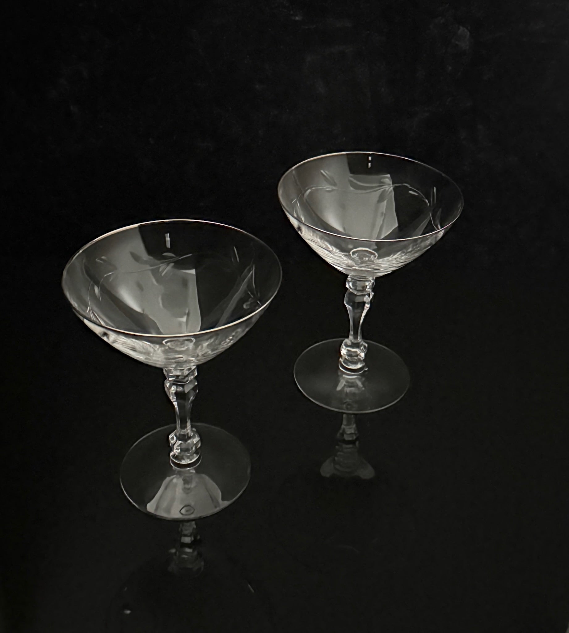 Two clear cut-glass coupe glasses with faceted stems and circular bases, photographed on a glossy black reflective surface against a black background.