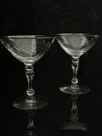 Two clear crystal coupe glasses with etched floral patterns, set against a dark reflective background.