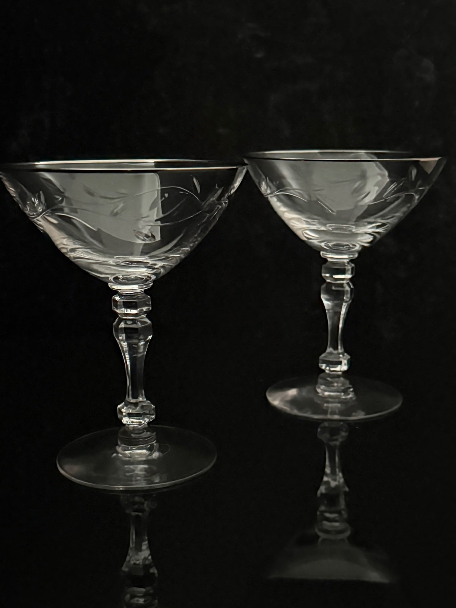 Two clear crystal coupe glasses with etched floral patterns, set against a dark reflective background.