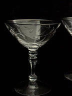 A clear cut-crystal coupe glass with a faceted stem and circular base, photographed against a black background. A second similar glass is partially visible on the right edge.