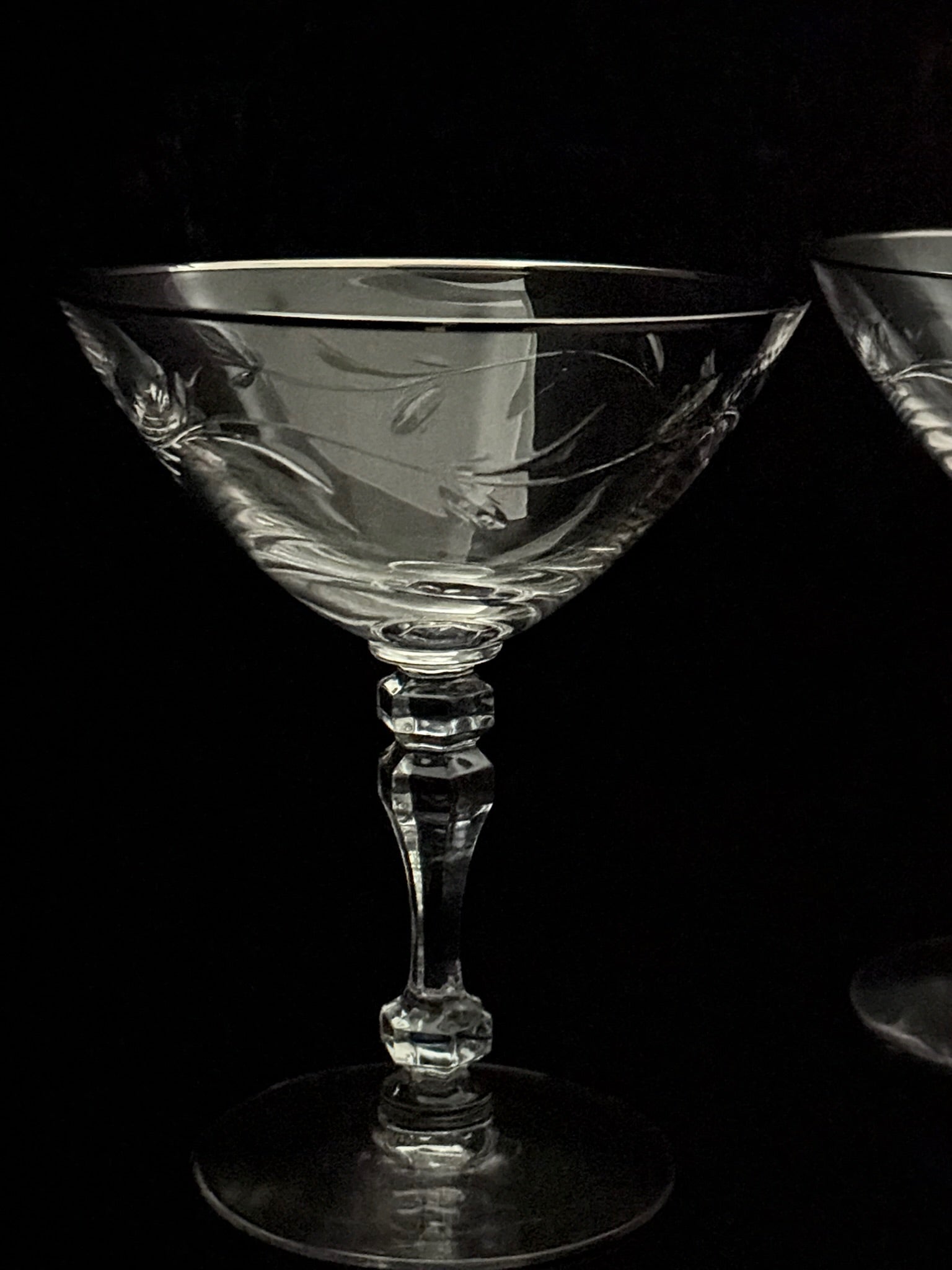 A clear cut-crystal coupe glass with a faceted stem and circular base, photographed against a black background. A second similar glass is partially visible on the right edge.