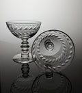 A clear cut-glass coupe glass and matching round plate with etched leaf patterns, displayed on a reflective dark surface against a light gray background.