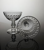 A clear cut-glass coupe glass and matching round plate with etched leaf patterns, displayed on a reflective dark surface against a light gray background.