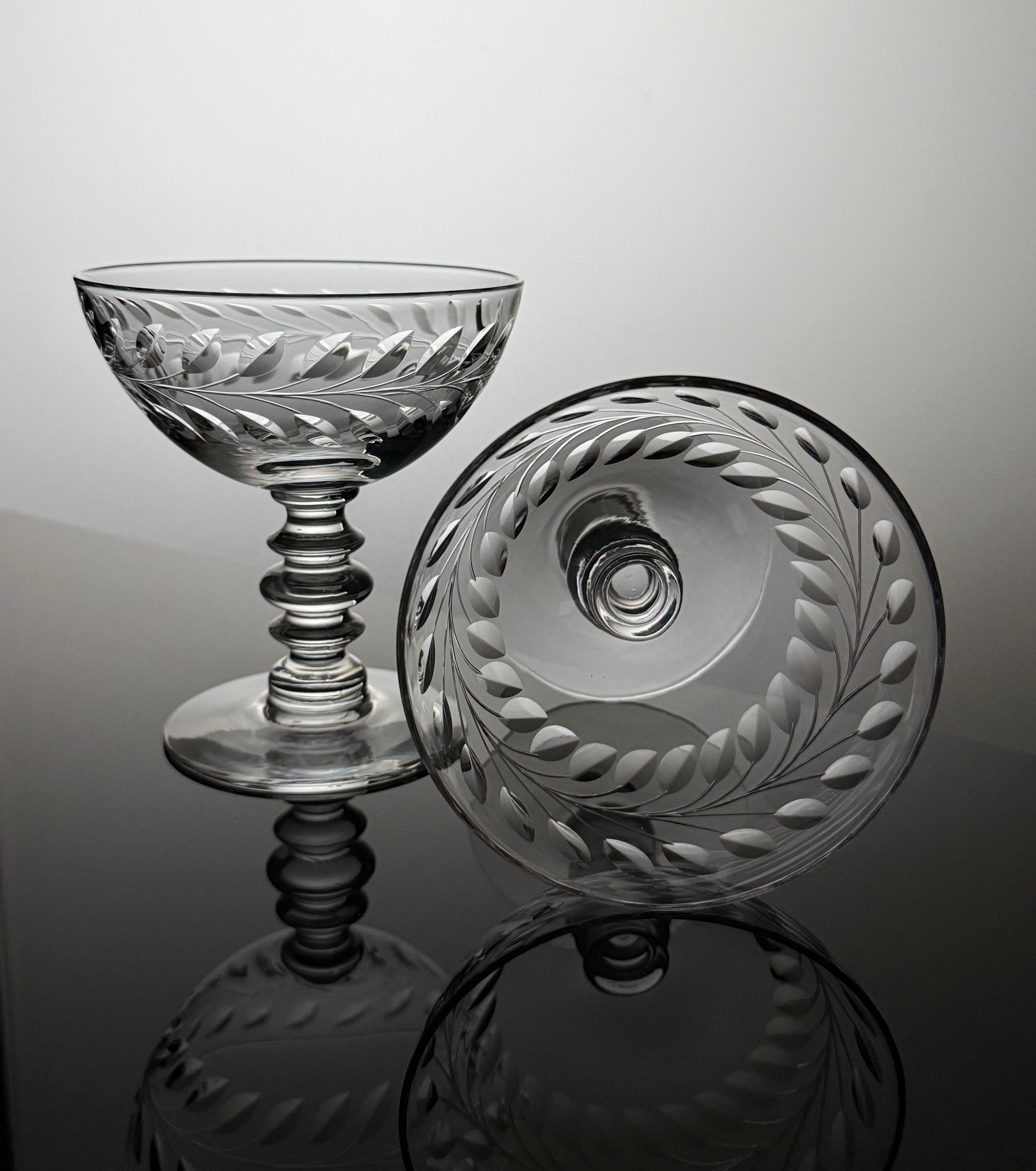A clear cut-glass coupe glass and matching round plate with etched leaf patterns, displayed on a reflective dark surface against a light gray background.