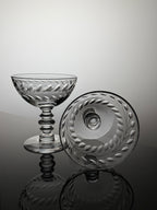 A clear cut-glass coupe glass and matching round lid with leafy border detailing, displayed on a reflective dark surface against a light gray studio background.