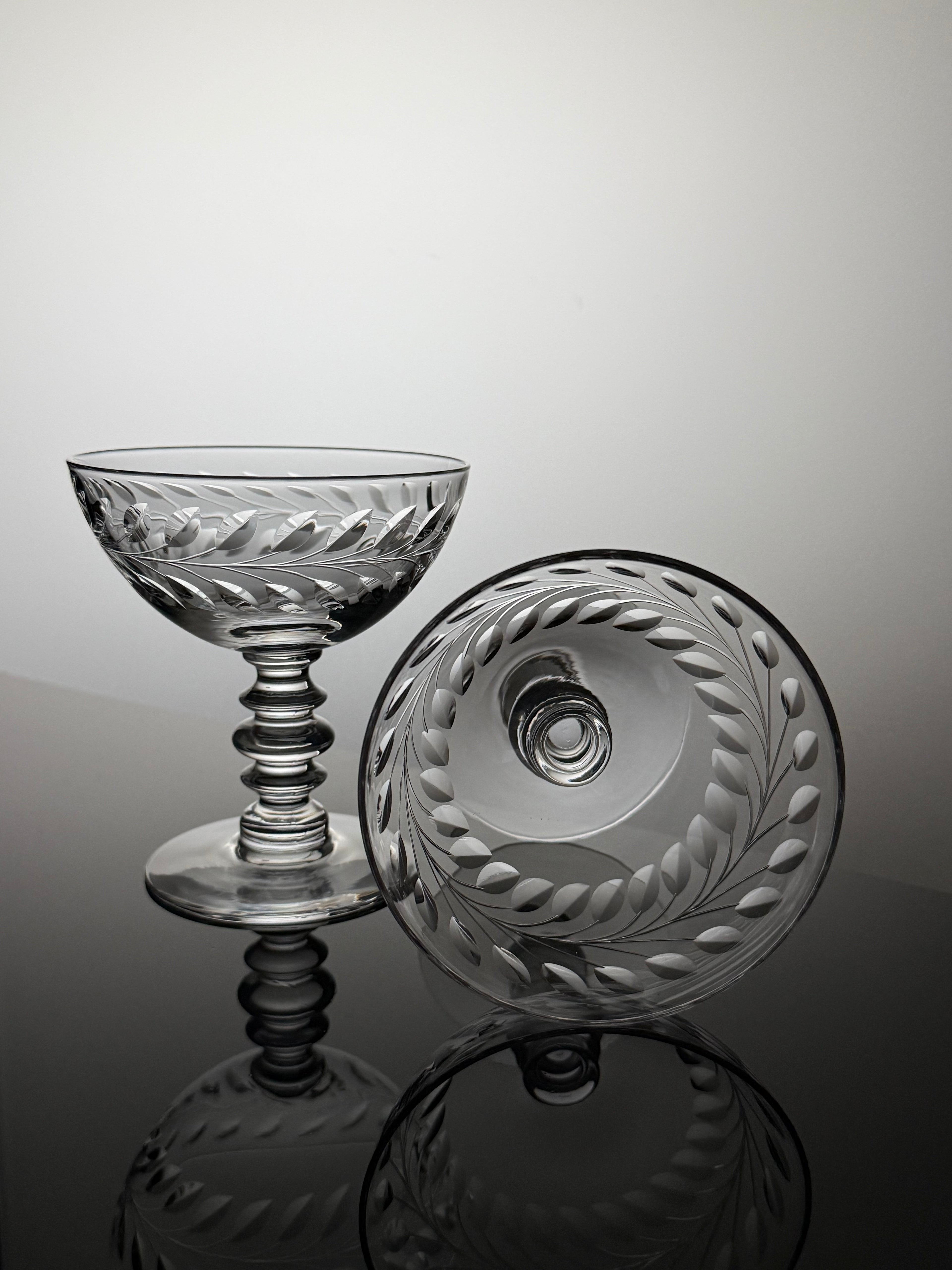A clear cut-glass coupe glass and matching round lid with leafy border detailing, displayed on a reflective dark surface against a light gray studio background.
