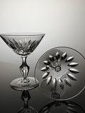 A clear cut-crystal coupe glass and its matching circular base plate, both featuring an intricate floral and geometric pattern, displayed on a reflective dark surface against a light gray background.