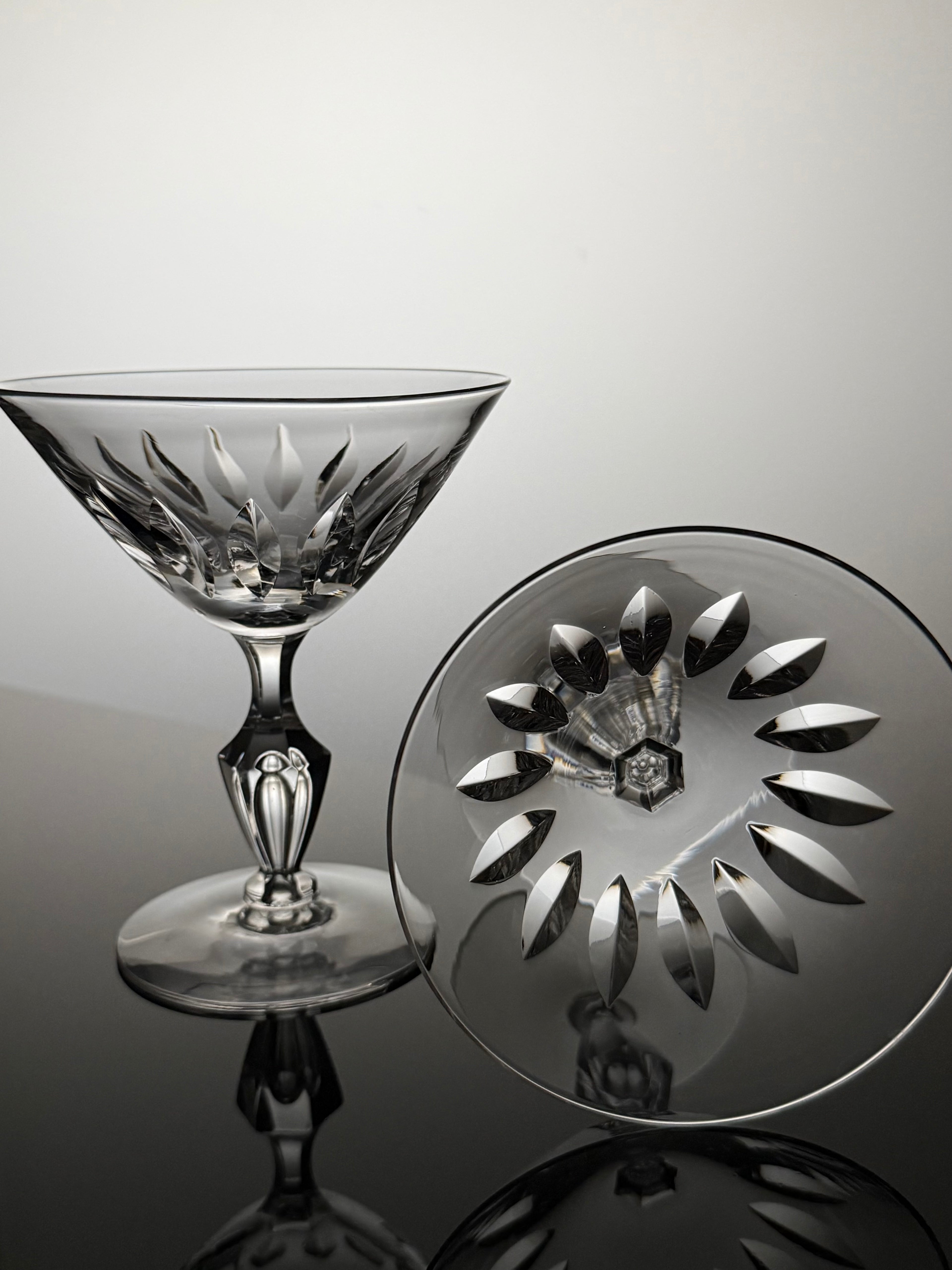 A clear cut-crystal coupe glass and its matching circular base plate, both featuring an intricate floral and geometric pattern, displayed on a reflective dark surface against a light gray background.