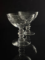 Two clear cut-crystal champagne coupes with flared stems, set against a dark reflective background.