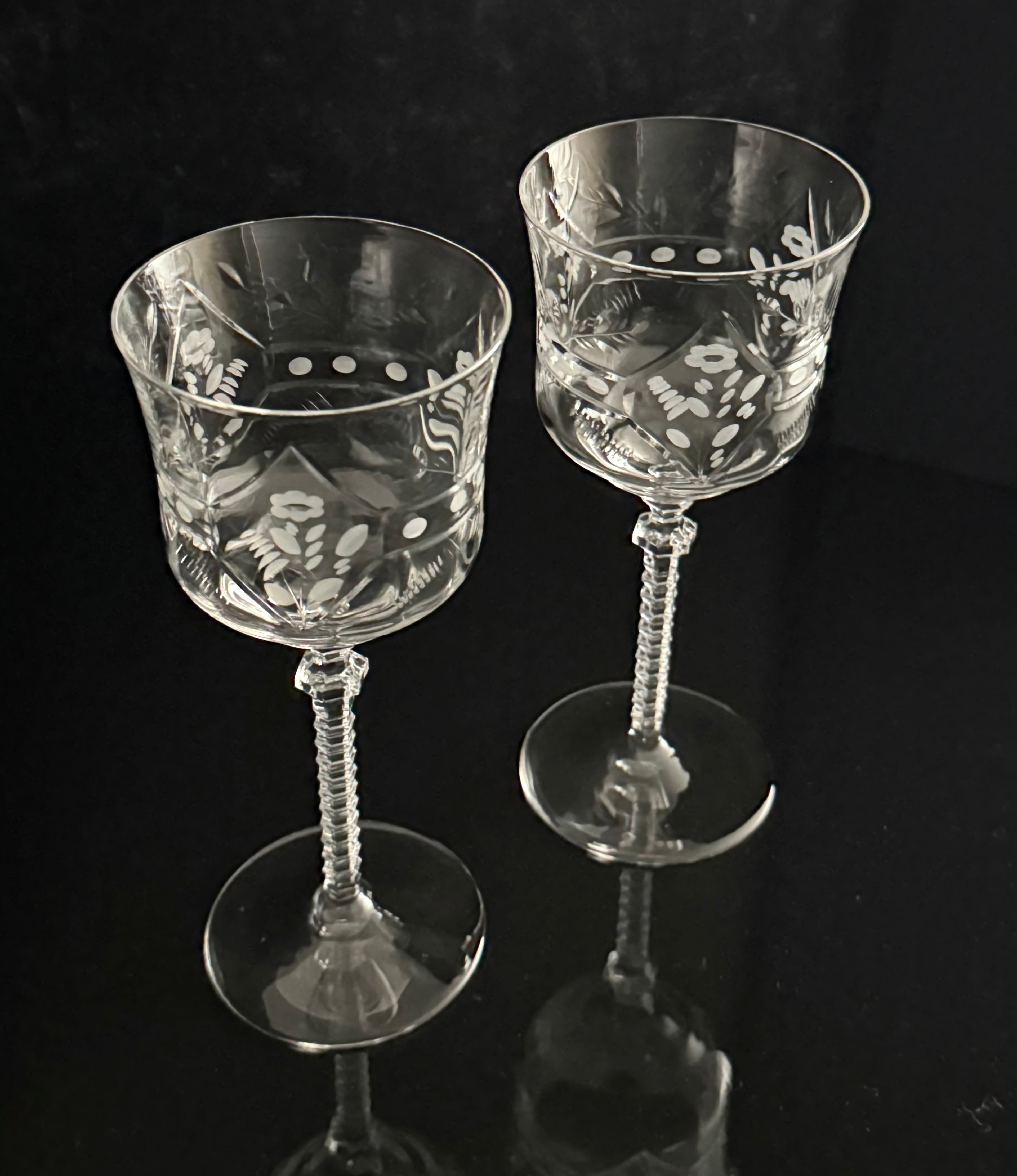 Two clear cut-glass goblets with ornate floral and geometric engravings, standing on a reflective black surface against a dark background.